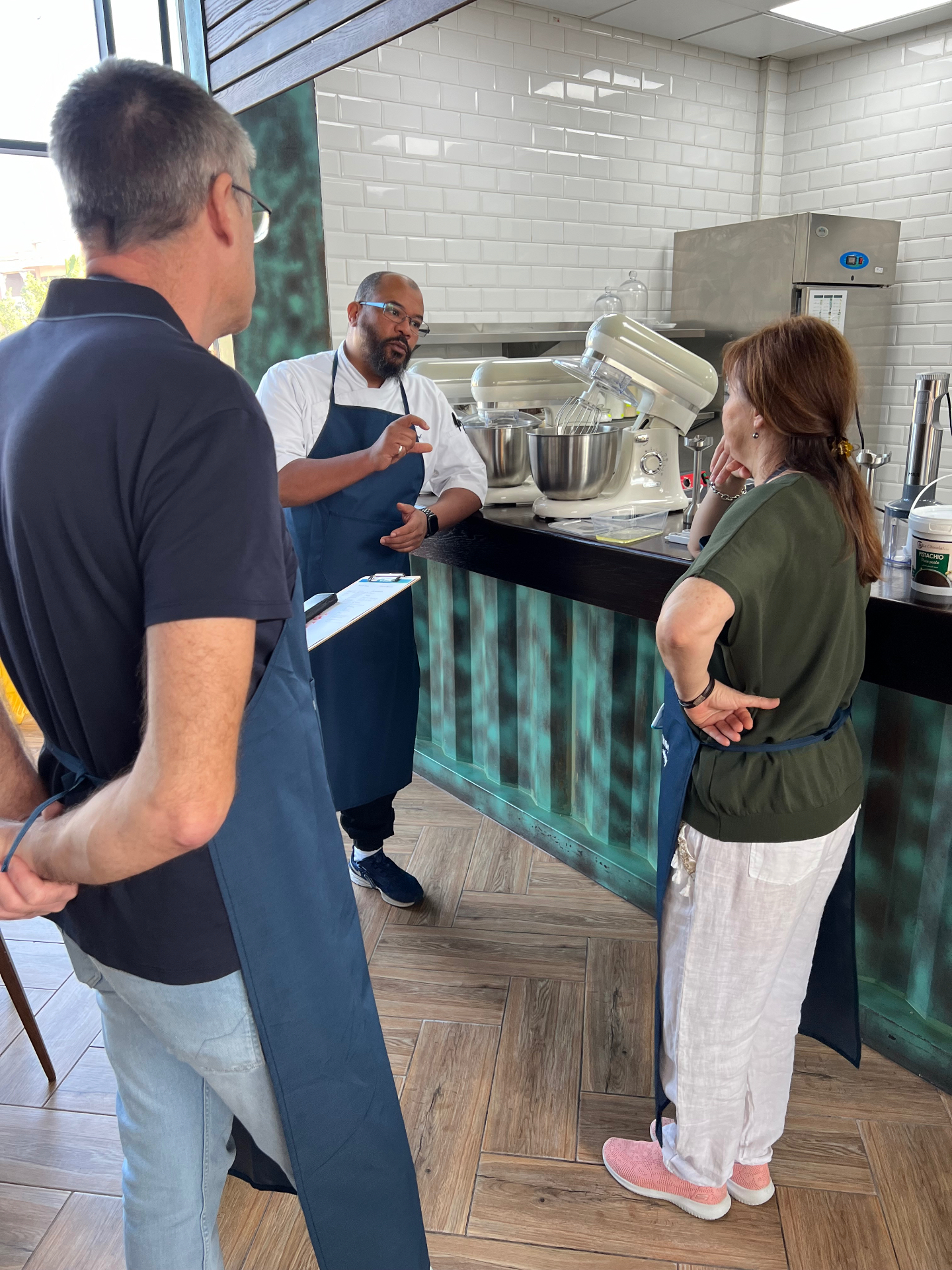 Chef Ahmed Badaoui expliquant une technique de pâtisserie à deux élèves en tablier dans un atelier culinaire professionnel
